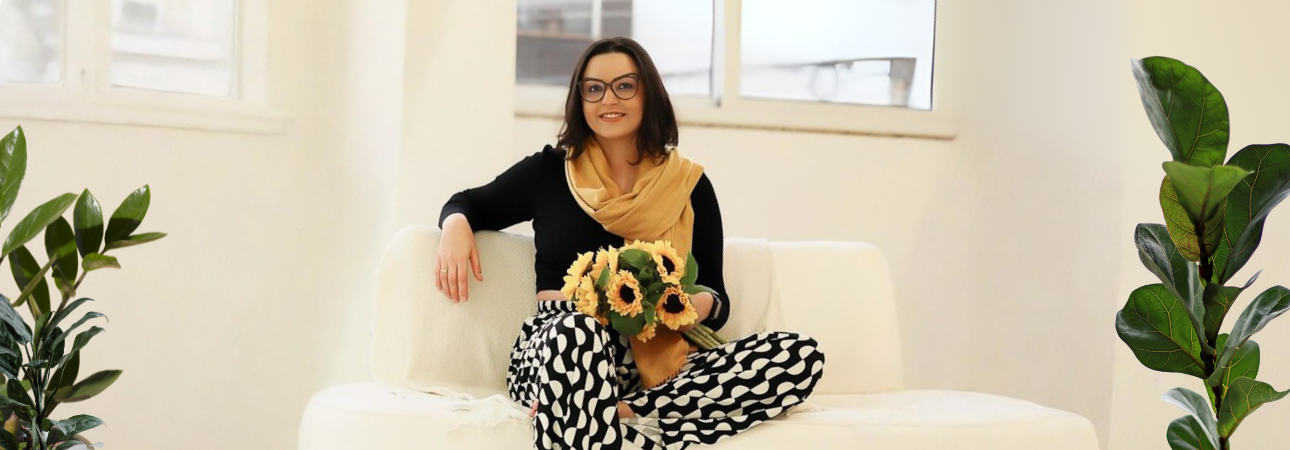 Natally Buzzatto, psicóloga, sentada em sofá claro com um buquê de girassóis.