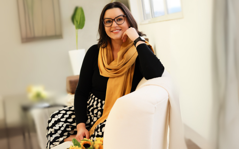 Natally Buzzatto, psicóloga, sentada em sofá claro, sorrindo, em ambiente de atendimento acolhedor.